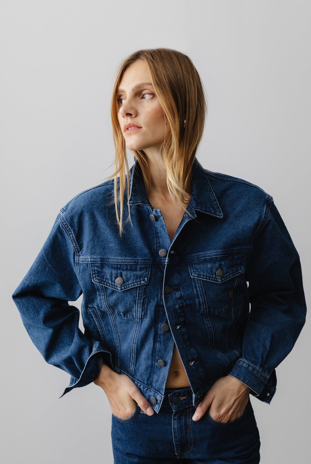 Casual denim jacket in deep indigo worn by a person with hands in pockets, featuring button details and a classic collar.