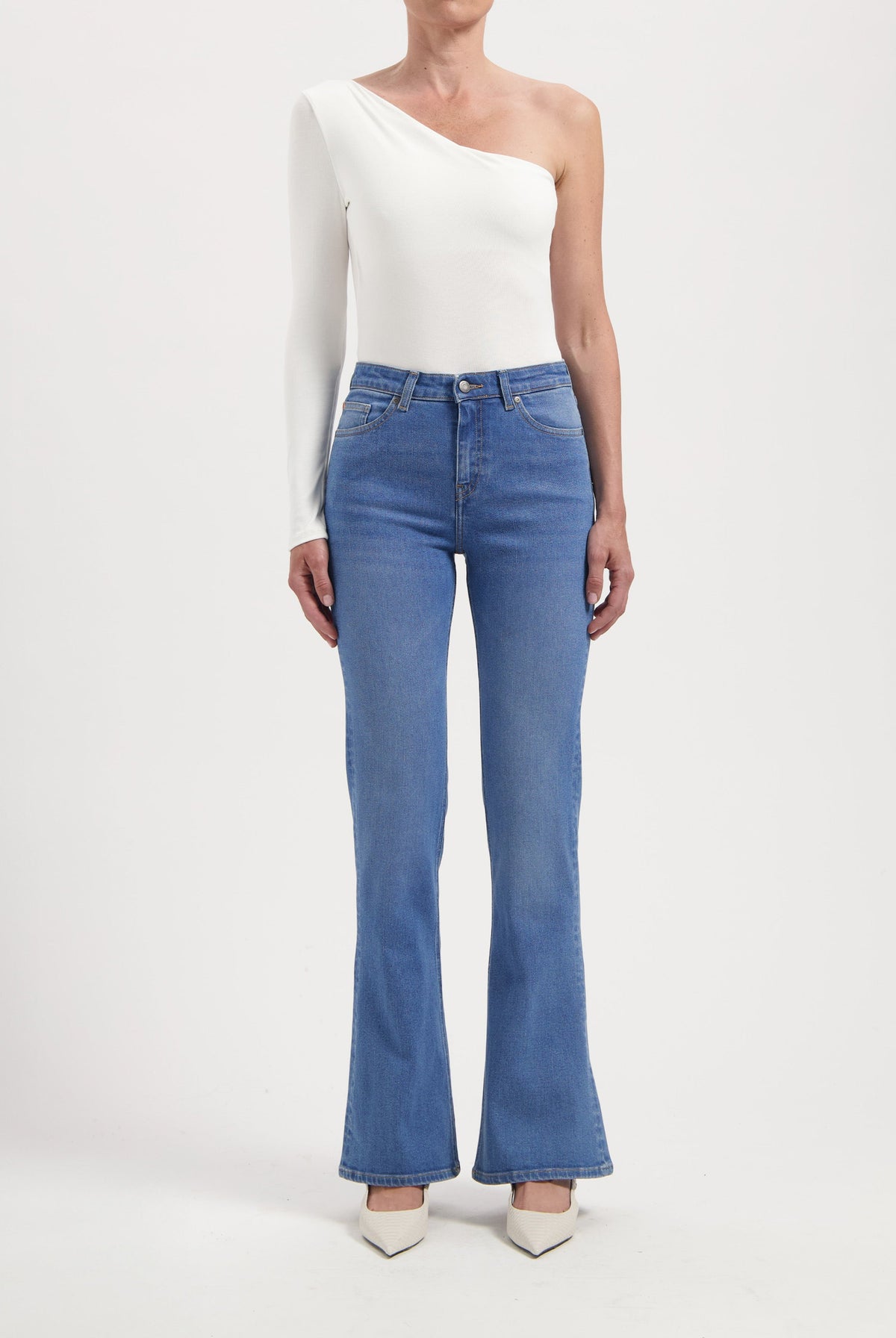 One-shoulder white top with long sleeve paired with high-waisted blue flare jeans, styled with white pointed heels.
