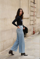 Woman wearing high-waisted light blue jeans, dark long-sleeve top, and black ankle boots on a city sidewalk.