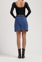 Back view of a woman in a black, long-sleeve top and a blue denim skirt, paired with sleek, black ankle boots.